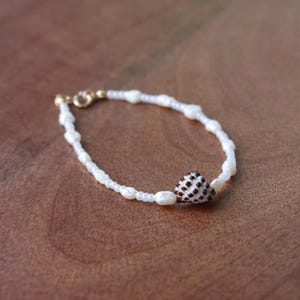 May include: A delicate bracelet featuring white pearls and a small seashell with brown spots. The bracelet has a gold clasp and is displayed on a wooden surface. This jewelry piece is perfect for adding a touch of coastal charm.