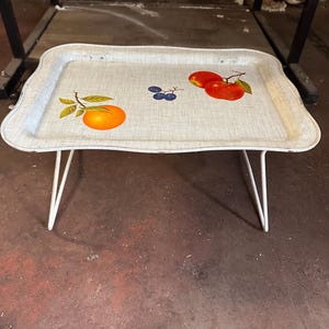 May include: A vintage metal tray table with a white frame and legs. The tray features a light beige background with illustrations of an orange, grapes, and tomatoes. The tray table is supported by white hairpin legs.