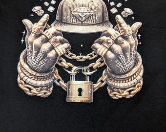 May include: Black t-shirt featuring a graphic of a diamond-studded baseball cap and hands adorned with rings and bracelets. A gold chain and padlock are also present in the design, along with scattered diamond shapes.