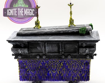 May include: A miniature coffin with a green hand reaching out. The coffin is gray with a purple and gold decorative drape. Two ornate candle holders with white candles sit on top. The logo for Ignite the Magic Design Co. is in the background.