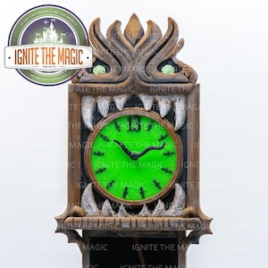 May include: A wooden wall clock with a green face and black hands. The clock is framed by a detailed, monster-like design with teeth and eyes. The text "IGNITE THE MAGIC" is visible on the clock face and the logo.