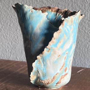 May include: A handmade ceramic vase with a unique, organic shape. The vase features a blend of light blue and white hues with brown accents along the rim. The textured surface adds to its artistic appeal. A decorative piece for home decor.