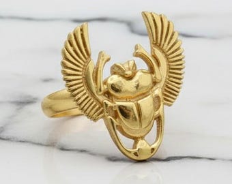 May include: A gold-coloured ring with a scarab beetle design and outstretched wings. The ring has a polished finish. The ring is set against a marble background, highlighting the intricate details and craftsmanship.
