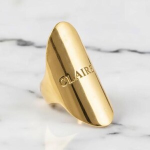 Może przedstawiać: Złoty pierścionek z wygrawerowanym imieniem "CLAIRE" z przodu. Pierścionek ma gładką, wypolerowaną powierzchnię i unikalny, wydłużony kształt. Pierścionek jest umieszczony na białym marmurowym tle.