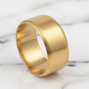 Op de afbeelding: Een goudkleurige ring met een brede band. De ring heeft een glad, gepolijst oppervlak en een eenvoudig ontwerp. De ring wordt getoond op een wit marmeren oppervlak.