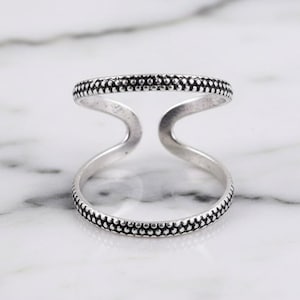 Può includere: Un anello a doppia fascia color argento con un motivo a pois lungo i bordi. L'anello ha un design aperto unico, con due fasce collegate da un ponte curvo. L'anello è esposto su una superficie di marmo bianco.