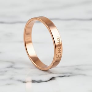 Puede incluir: Anillo de oro rosa con el nombre "Emma" grabado en el lateral. El anillo está sobre un fondo de mármol. El anillo tiene un diseño de banda simple.