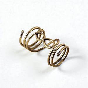 May include: A gold-toned ring with an infinity symbol design. The ring features multiple coiled bands and a unique, open design. The metal has a slightly aged appearance, adding to its vintage appeal. The ring is set against a white background.