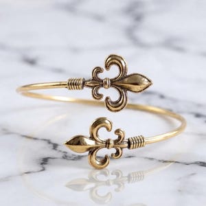 May include: A gold-toned metal cuff bracelet featuring two symmetrical fleur-de-lis motifs at each open end. The band has a smooth, polished finish with ribbed detailing near the decorative fleur-de-lis elements, creating a continuous open loop.