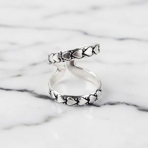Puede incluir: Un anillo plateado con un diseño de doble banda. Las bandas están conectadas en la parte superior e inferior, y cada banda está adornada con un patrón de corazones repetidos. El anillo se muestra sobre una superficie de mármol blanco y gris.