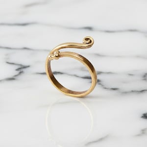 Könnte beinhalten: Ein goldfarbener Ring mit einem einzigartigen Design. Der Ring hat ein geschwungenes Band, das den Finger umschließt, mit einem kleinen floralen Detail und einer Wirbelverzierung an der Spitze. Der Ring liegt auf einer weißen Marmoroberfläche.