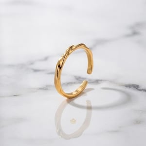 May include: A gold-toned open ring featuring a twisted, textured band design. The ring has a polished metallic finish and a gap at the top, creating a continuous, fluid shape that catches the light across its curved, uneven surface.