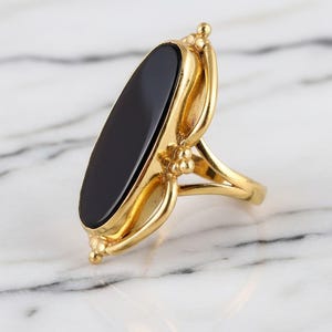 May include: A gold-colored ring featuring a large, oval-shaped black stone. The ring has an ornate design with decorative elements and a polished finish. The ring is set against a marble background.