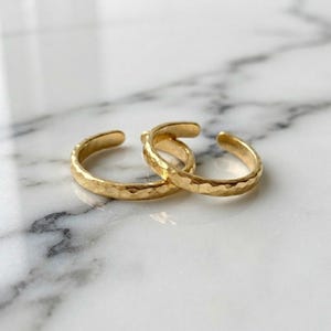 Könnte beinhalten: Zwei goldfarbene Ringe mit offenem Design und einer gehämmerten, strukturierten Oberfläche. Die Ringe haben ein poliertes Finish, das Licht reflektiert, und zeigen eine einfache, kreisförmige Bandstruktur mit einer kleinen Lücke an der Öffnung zur Anpassung.