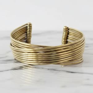 Puede incluir: Un brazalete de puño dorado con múltiples bandas finas y paralelas que se cruzan. Los extremos del brazalete tienen una serie de barras verticales y redondeadas. El brazalete se exhibe sobre una superficie de mármol blanco y gris.