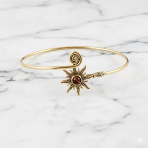 Puede incluir: Un brazalete de puño dorado con un diseño de sol y una piedra central marrón. El brazalete tiene un detalle en espiral y acentos decorativos. La joya se exhibe sobre una superficie de mármol blanco.