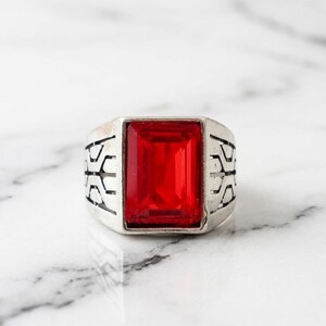 May include: A silver-toned ring with a large, rectangular, red gemstone. The band features a geometric design on each side of the gemstone, creating a bold and eye-catching accessory.