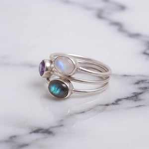 May include: A stack of three silver rings featuring oval-shaped gemstones. The top ring has a translucent white moonstone, the middle ring has a small purple amethyst, and the bottom ring has a dark grey labradorite with blue iridescence.