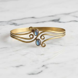 May include: A gold-coloured metal cuff bracelet with two oval, blue gemstones. The bracelet has a three-band design with decorative swirls on each side of the gemstones. The bracelet is displayed on a white marble surface.