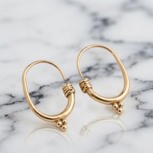 May include: A pair of gold hoop earrings with an oval shape. Each earring features a textured band section with horizontal ridges and a small cluster of three tiny spheres at the base of the hoop, creating a detailed and polished metallic finish.