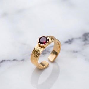 Puede incluir: Un anillo de oro con una piedra preciosa redonda de color rojo oscuro engastada en un bisel. La banda tiene un diseño abierto y un acabado martillado texturizado en toda su superficie. El anillo está colocado en posición vertical.