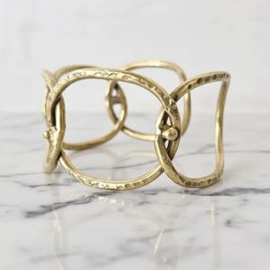 May include: A gold-colored metal cuff bracelet with an open, interlocking design. The bracelet features a series of connected oval and rectangular shapes, creating a unique and stylish accessory. The metal has a textured surface.
