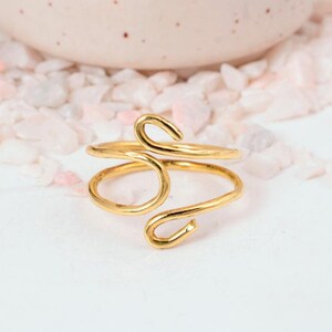 May include: A gold-colored ring with a unique, open design. The ring features a swirling, abstract shape with curved lines and a polished finish. The ring is set against a white surface with pink and white decorative elements in the background.