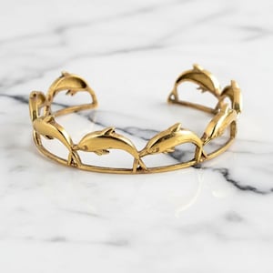 May include: A gold-toned metal cuff bracelet featuring a repeating openwork design of leaping dolphins connected by a thin circular band. The bracelet has a polished finish and an open back, showcasing a series of stylized dolphin figures.