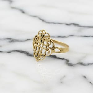 Puede incluir: Un anillo dorado con un diseño calado ornamentado. El anillo presenta un patrón central de filigrana con detalles en espiral y una banda lisa. El anillo está sobre una superficie de mármol blanco y gris.