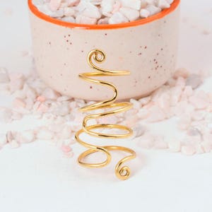 May include: A gold-colored, spiral-shaped ring. The ring has a unique design with a coiled central section and curved ends. The ring is displayed on a white surface with a decorative pot in the background filled with small, light-colored stones.