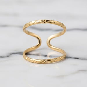 May include: A gold-colored double-finger ring. The ring features a delicate, etched pattern along the bands. The design curves gracefully, creating a unique and stylish accessory. The ring is displayed on a marble surface.