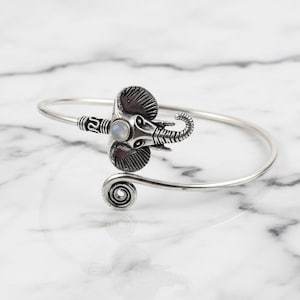 May include: Silver-toned elephant bracelet with a curved design. The elephant head features detailed tusks, ears, and a small, round, light-colored stone. The bracelet has a spiral detail at the end and is displayed on a marble surface.