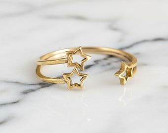 May include: A gold-coloured open ring with three star-shaped cutouts. The ring has a double band design and is set against a white and grey marble surface. The stars are evenly spaced, adding a celestial touch to the jewellery.