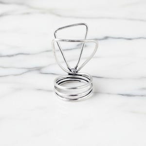 Puede incluir: Un soporte de anillo de metal plateado con una parte superior triangular y una base de tres anillos circulares. El soporte está diseñado para exhibir artículos y se encuentra sobre una superficie de mármol blanco.