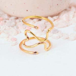 May include: A gold-colored, abstract ring with a unique, flowing design. The ring features multiple intertwined loops and curves, creating an eye-catching, modern aesthetic. The ring is displayed on a white surface with small, light-colored stones in the background.