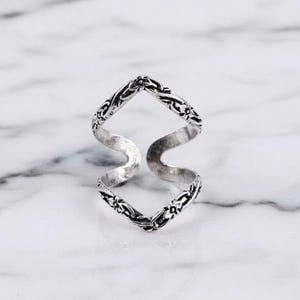 May include: A silver ring with an open, double-S design. The ring has an intricate floral pattern along the edges. The ring is displayed on a white marble surface.