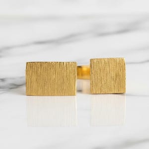 Puede incluir: Un par de gemelos dorados con un diseño rectangular texturizado. Los gemelos se muestran sobre una superficie de mármol blanco, reflejando la luz. El diseño presenta un efecto de metal cepillado.