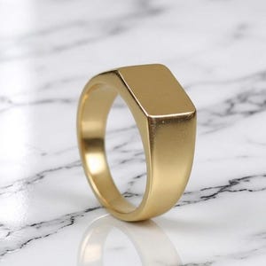 May include: A gold signet ring with a rectangular face. The ring has a smooth, matte finish and a classic, minimalist design. The ring is shown on a white and grey marble surface.