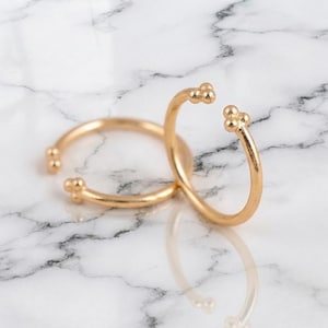 May include: Two gold-toned open rings featuring a smooth, thin band design. Each end of the rings is finished with a cluster of three small, rounded spheres, creating a delicate and symmetrical appearance for these adjustable jewelry pieces.