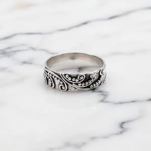 May include: A silver ring with an intricate floral pattern. The ring's design includes swirling lines and small dots, giving it a textured look. The ring is displayed on a white marble surface.