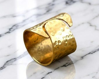 May include: A wide, gold-colored ring with a hammered texture. The ring has a unique design with an overlapping section. The ring is displayed on a white and gray marble surface.