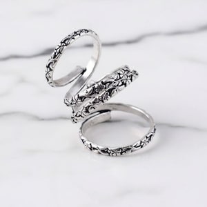Puede incluir: Un anillo ajustable de color plateado con un diseño floral. El anillo se enrolla alrededor del dedo en espiral, con tres bandas conectadas. El detalle floral es consistente en todas las bandas.