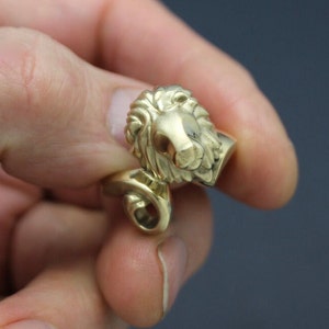 May include: A gold-colored lion head pendant with detailed mane and facial features. The pendant has a loop for attaching to a chain or other jewelry. The lion's head is facing to the right.