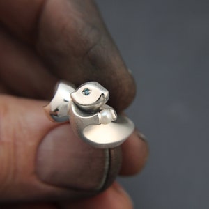 May include: A silver ring featuring a stylized squirrel design. The squirrel has a small blue gemstone eye and is holding a small pearl. The ring is held by a person's fingers against a dark background.