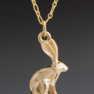May include: A gold-colored necklace featuring a pendant in the shape of a rabbit. The rabbit pendant is detailed and has long ears. The chain is also gold-colored and has an oval link design. The background is a dark gray.
