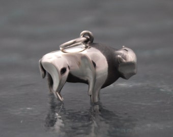 May include: A polished silver buffalo charm with a loop for attaching to a necklace or bracelet. The charm has a detailed design, with a dark grey head and a shiny silver body. The buffalo is standing, and the charm is likely a pendant.