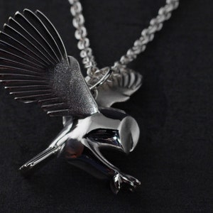 May include: A silver-toned pendant necklace featuring a stylized bird in flight. The bird has outstretched wings with detailed feathering and a polished, reflective body. The necklace chain is a textured, linked design.