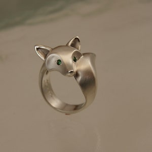 May include: A silver fox ring with green gemstone eyes. The ring features a detailed fox head design with a curved tail forming the band. The fox's eyes are small, round, and a vibrant green color. The ring is made of a matte silver metal.