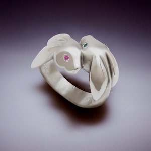 May include: A silver ring with a unique design featuring two stylized rabbit heads facing each other. One rabbit has a pink gemstone eye, the other has a blue gemstone eye. The ring is set against a gradient background.