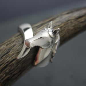 May include: A silver-colored ring featuring a horse head design. The ring wraps around the finger, with the horse's head prominently displayed. The horse has a detailed mane and a pointed ear. The ring is made of a shiny, reflective metal.